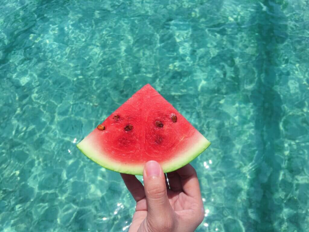 Hand holding a slice of watermelon over a pool.